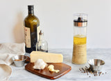 A marble countertop with a bottle of olive oil, a small glass vinegar bottle, a block of cheese, garlic cloves, a dressing shaker filled with vinaigrette, two 6 oz stainless steel sauce cups with spices, and scattered black peppercorns. A beige napkin sits on the left.