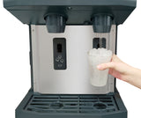 A hand holds a clear cup filled with ice under the water dispenser of a black and stainless steel ice and water dispenser. Water pours into the cup from two nozzles, while an "ICE" button and drip tray are visible on this touchfree infrared dispensing machine.