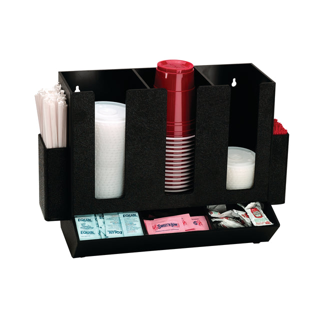 A black countertop condiment organizer holds white stirrers, clear plastic lids, red plastic cups, and red straws. The bottom section of this cup organizer stores sugar, sweetener, and condiment packets in neat compartments against a white background.
