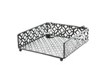A black powder coated metal napkin holder from the TableCraft Farmhouse Collection features a geometric cut-out pattern, four round feet, and a weighted arm with a ball end to keep napkins in place. Photographed on a white background.