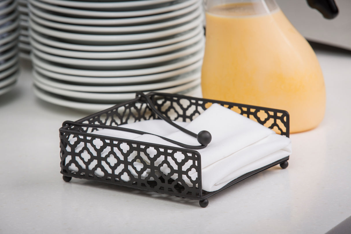 A black powder coated metal Farmhouse Collection Napkin Holder by TableCraft with a geometric lattice design holds white napkins on a countertop, set against a stack of plates and a jug of orange juice in a cozy breakfast kitchen.