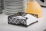 A black powder coated metal Farmhouse Collection Napkin Holder by TableCraft with a geometric lattice design holds white napkins on a countertop, set against a stack of plates and a jug of orange juice in a cozy breakfast kitchen.