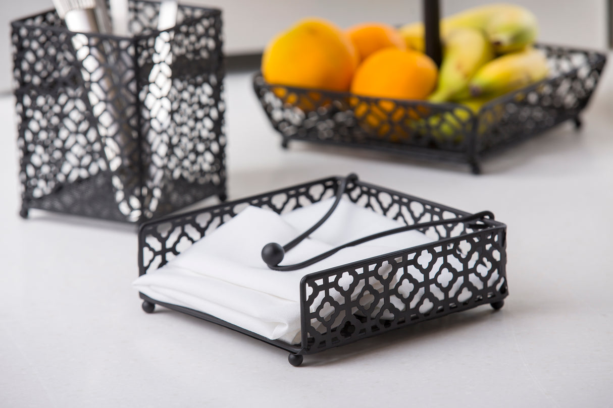 A decorative black powder coated metal Farmhouse Collection Napkin Holder by TableCraft holds white napkins and a metal weight. In the background, matching metal containers hold cutlery and assorted fruits on a white countertop.