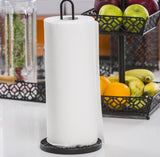 A roll of white paper towels stands upright on a black metal paper towel holder atop the kitchen counter. In the background, a two-tiered fruit basket holds apples, bananas, and oranges, with a clear vegetable container partially visible.