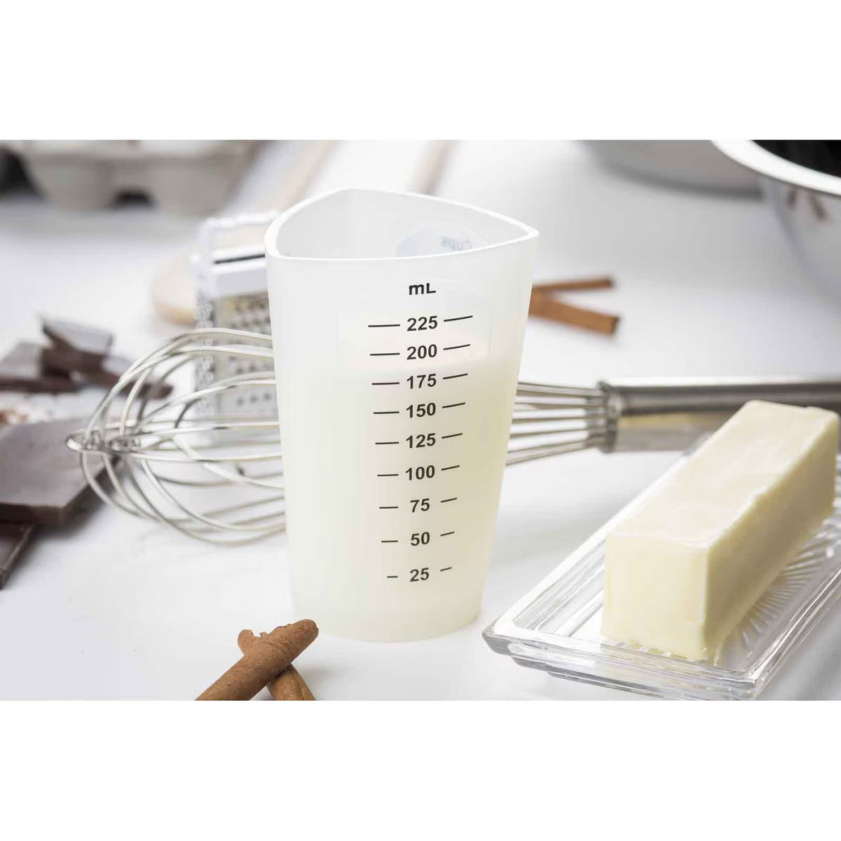 A dishwasher safe plastic measuring cup marked in milliliters sits on a kitchen counter. Behind it are a metal whisk, baking ingredients, a stick of butter on a glass dish, cinnamon sticks, chocolate pieces, and eggs in a carton.