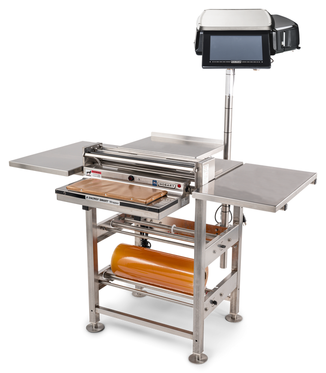 A commercial stainless steel wrapping machine with a mounted touchscreen, roll film capacity, side shelves, heated seal plate, and cutting tray. The device stands on four legs and is designed for efficient packaging of food or products.