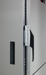 Close-up of a stainless steel cabinet door on a commercial mobile food cabinet, showing a black recessed handle on the left and a silver and black latch handle on the edge, secured with screws, with visible hinges and textured finish.