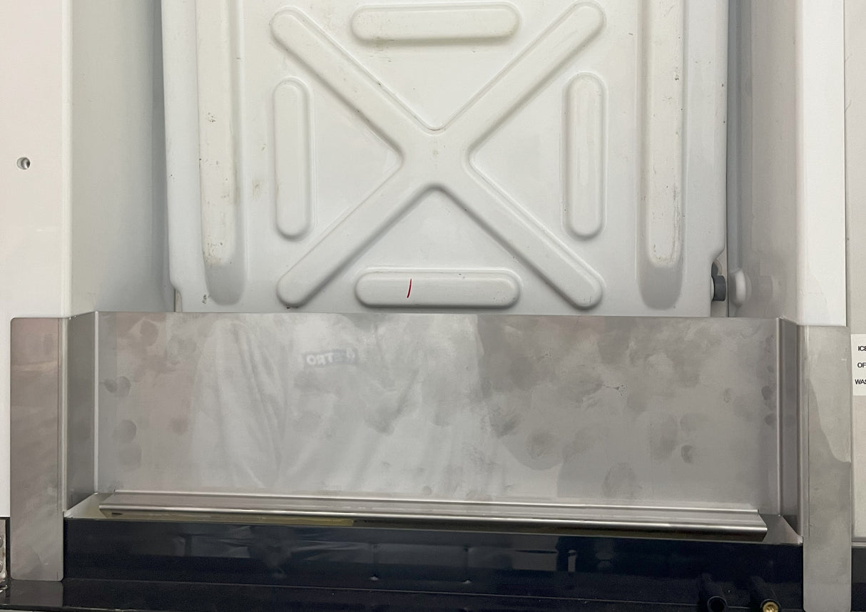 A large white metal panel with an embossed X pattern, possibly part of a beverage dispenser deflector, is mounted above a stainless steel shelf or counter. The reflective surface of the shelf shows faint reflections of people and surroundings.
