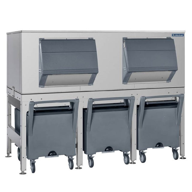 A large Scotsman industrial ice storage bin system with a stainless steel body, two front flip-up doors, and three gray bins on wheels below for collecting ice; the bins have handles, making this an efficient ice transport system on adjustable legs.