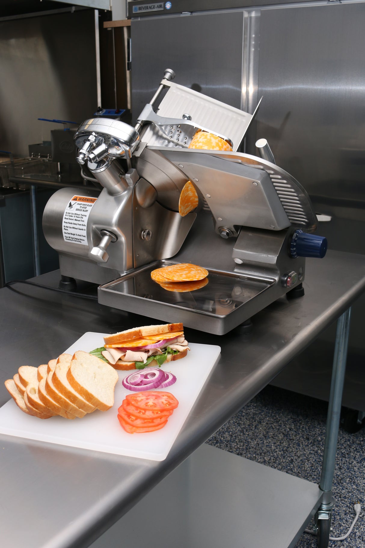 A Globe GC512 compact slicer sits on a stainless steel counter with a sliced loaf and deli meat. In the foreground, a cutting board holds bread, tomato, red onion, lettuce, and cheese—ingredients ready for making sandwiches.