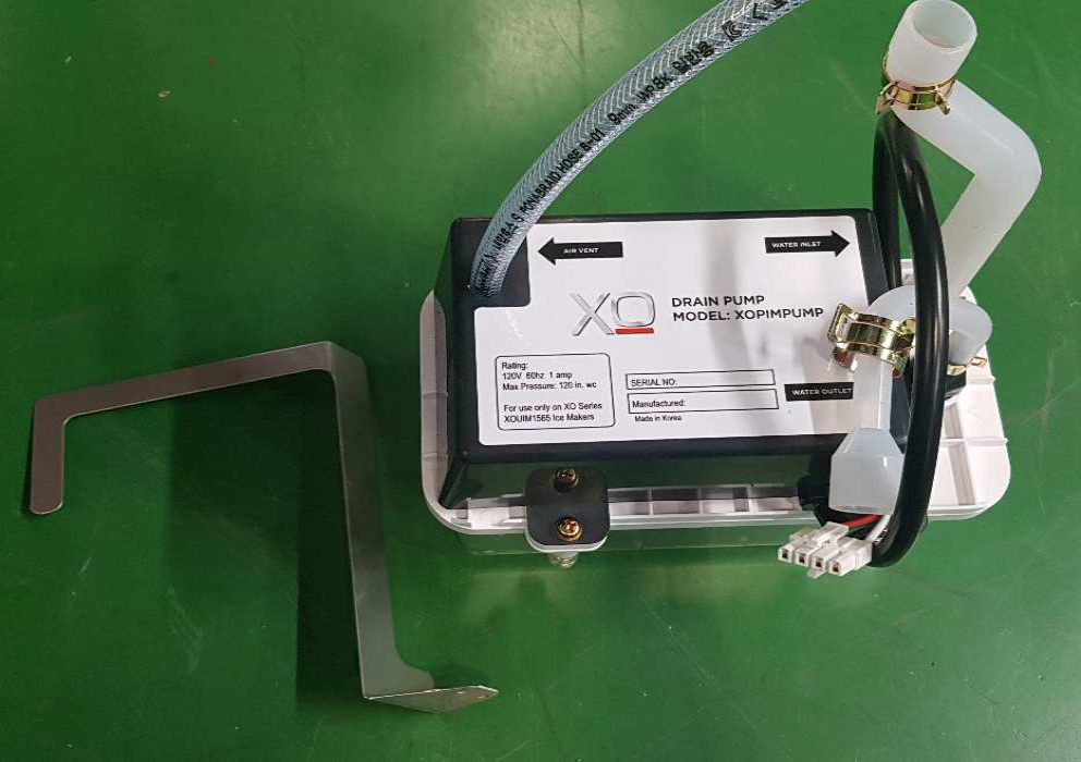 A small Icetro America XQ drain pump unit labeled “MODEL: XQMINPUMP” sits on a green surface. Attached tubes and wires connect to a white plastic container. The Drain Pump Kit’s metal mounting bracket, IU-0090-AN, is placed next to it on the left.