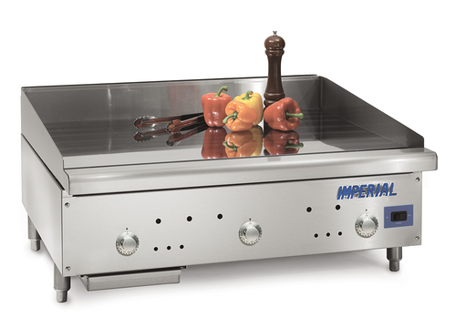 A stainless steel countertop gas griddle with three control knobs and the Imperial brand on the front. On top, metal tongs, three bell peppers, and a tall pepper grinder rest on its clean, reflective surface. Features a solid state thermostat for precision.