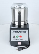 A Robot Coupe R301U.D commercial food processor is shown upright on a white surface. The stainless steel bowl has a clear lid, ideal for efficient vegetable prep in busy kitchens. The control panel displays green and red power buttons, model information, and a 3.7-liter capacity. The power cord with plug is visible.