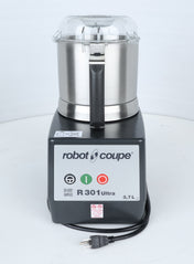 A Robot Coupe R301U.D commercial food processor is shown upright on a white surface. The stainless steel bowl has a clear lid, ideal for efficient vegetable prep in busy kitchens. The control panel displays green and red power buttons, model information, and a 3.7-liter capacity. The power cord with plug is visible.