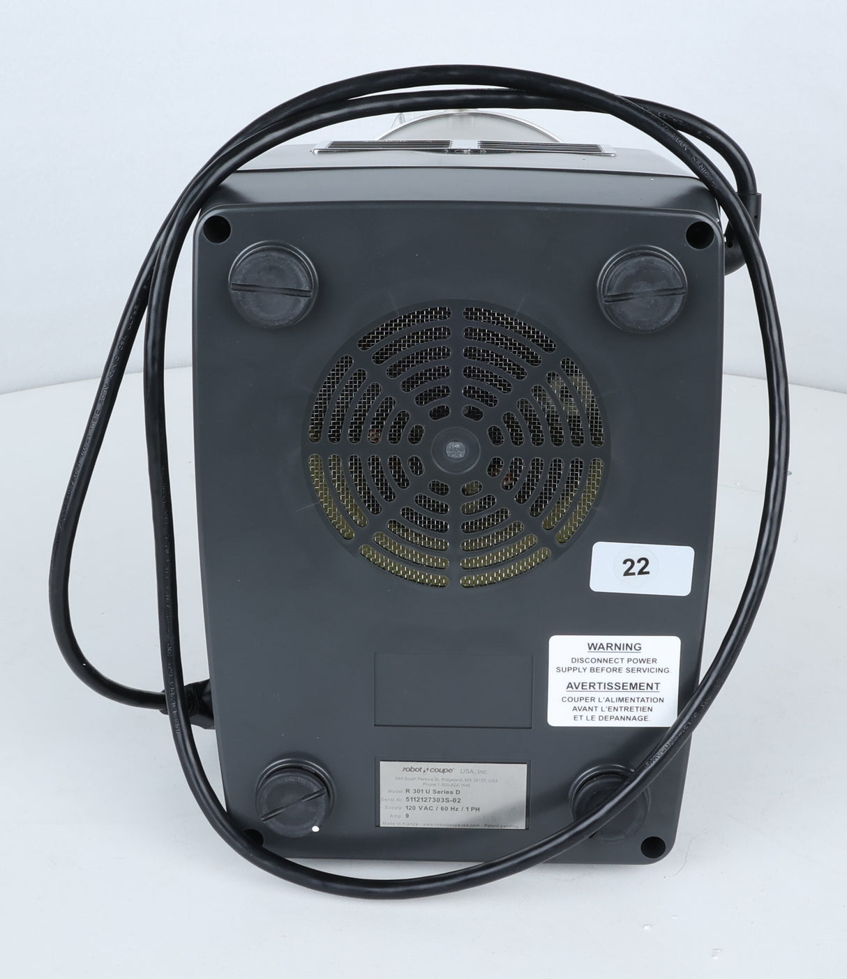 Bottom view of a black Robot Coupe R301U.D commercial food processor with four rubber feet, a circular vent with metal mesh, a coiled power cord, and several white labels, including a warning and number 22. The background is plain and light gray—ideal for efficient vegetable prep in any professional kitchen.