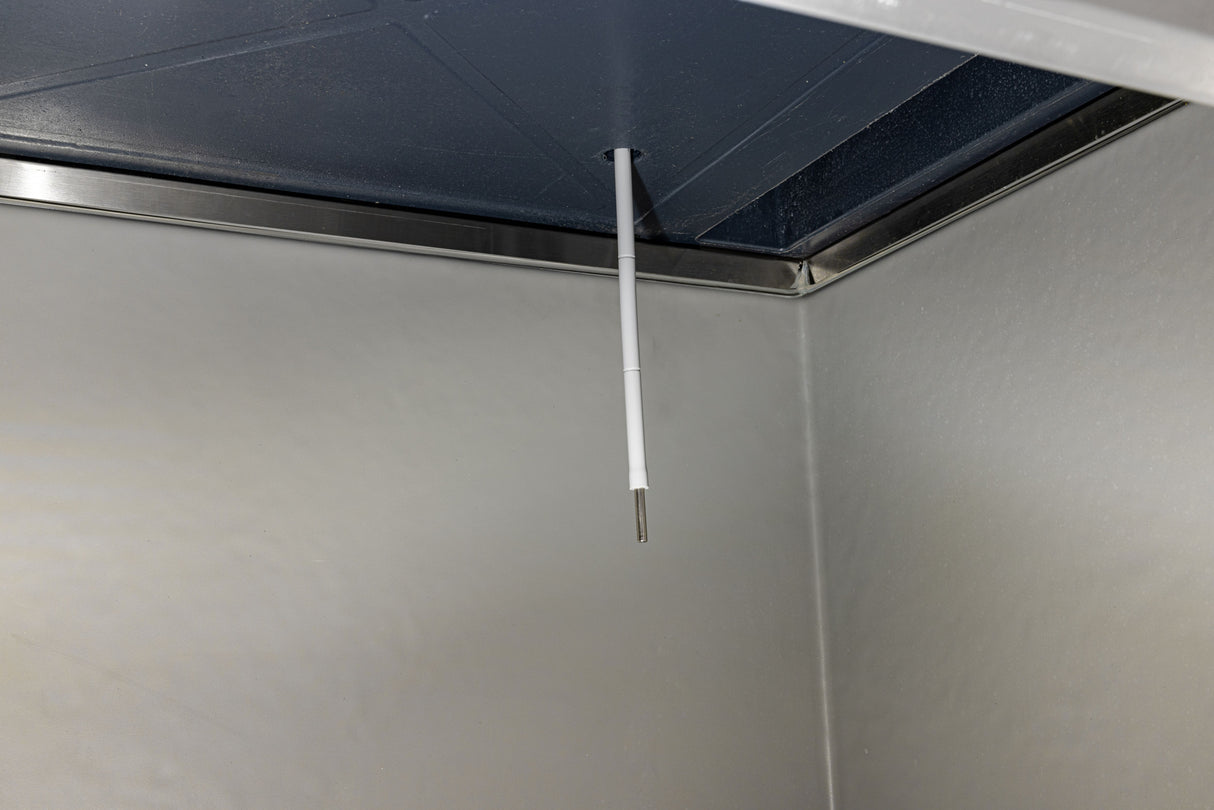 A white temperature probe using thermistor technology hangs from the ceiling inside a metallic, industrial-looking chamber with smooth, gray walls and a dark blue ceiling panel. The thin, cylindrical sensor extends downward near the corner of the chamber.