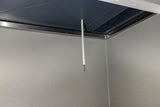 A white temperature probe using thermistor technology hangs from the ceiling inside a metallic, industrial-looking chamber with smooth, gray walls and a dark blue ceiling panel. The thin, cylindrical sensor extends downward near the corner of the chamber.