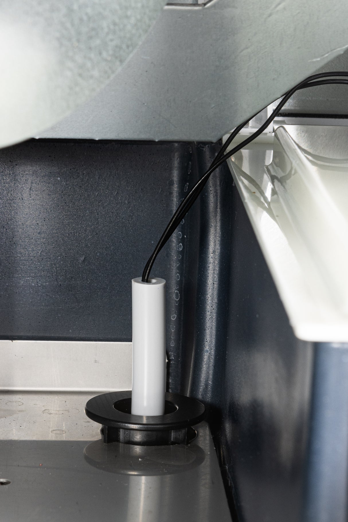 A close-up view of a white cylindrical probe featuring thermistor technology with black wiring, inserted vertically through a circular black mount into a metal panel, near a white tray inside industrial or laboratory equipment housing.