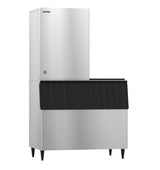 A Hoshizaki cube-style ice maker features a tall, rectangular stainless steel top unit for ice making and a large, matching bottom bin with a ribbed black lid for storage, all standing on four short black legs against a white background.