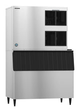 A commercial Hoshizaki ice maker with a stainless steel finish features a large rectangular shape, black vent panels on the upper right, a blue logo on the lower left, and a cube-style ice storage bin below with angled grooves. The unit stands on four short legs.