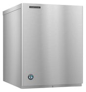 A commercial Hoshizaki Cube-Style ice maker with a plain front, boxy shape, and smooth surface. The Hoshizaki brand label sits on the top right, with a small round certification sticker near the bottom left corner. The unit rests on a flat surface.
