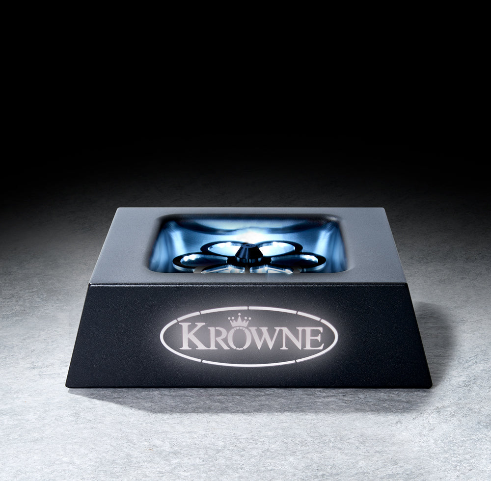 A square black metal glass chiller with stainless steel construction features a central circular rinser and four spokes, branded with a glowing “KROWNE” logo on the front. The illuminated rinser area highlights its sleek, industrial design.