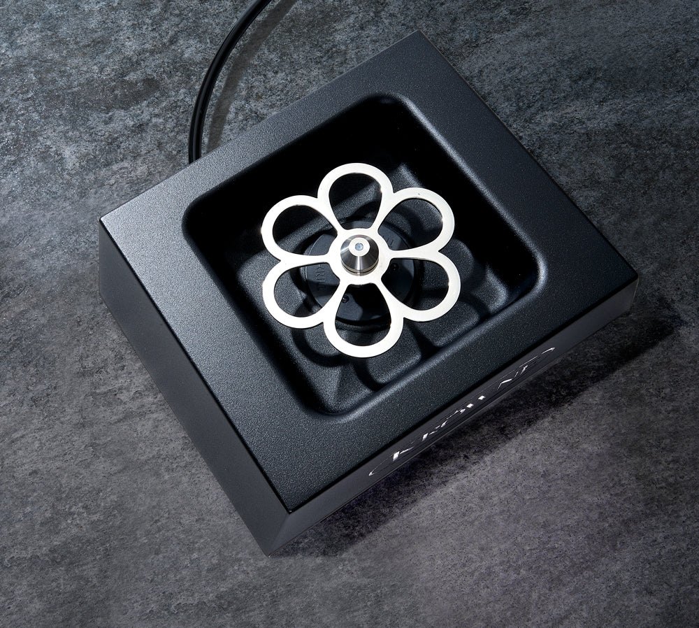 A top-down view of a black square Countertop Liquid CO2 Glass Chiller with a silver, flower-shaped stainless steel center mounted on a spindle. The device rests on a dark stone surface, with a power cord extending from the top.