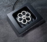A top-down view of a black square Countertop Liquid CO2 Glass Chiller with a silver, flower-shaped stainless steel center mounted on a spindle. The device rests on a dark stone surface, with a power cord extending from the top.