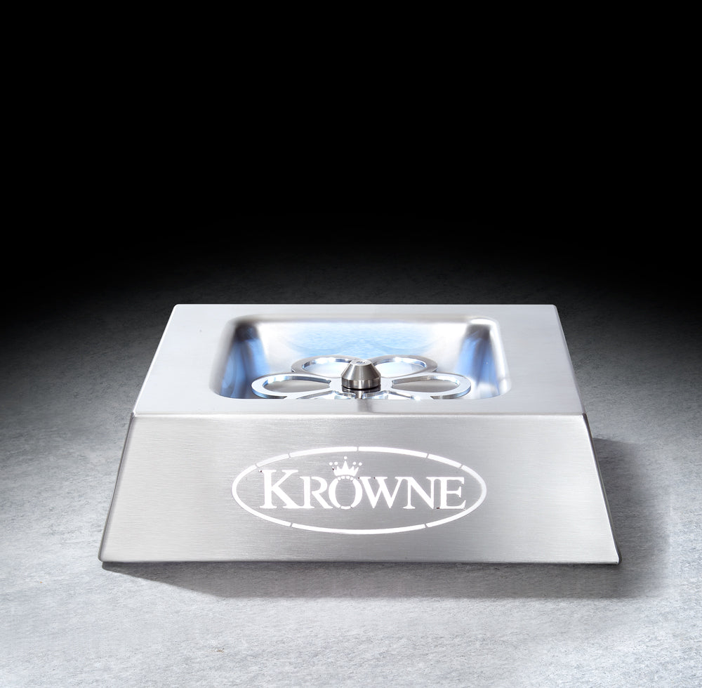 A stainless steel bar glass rinser with a square design, sloped front, and central spray nozzle—ideal for any setup using a countertop glass chiller—features the KROWNE name and crown logo on its front. It sits on a dark, textured surface against a black background.