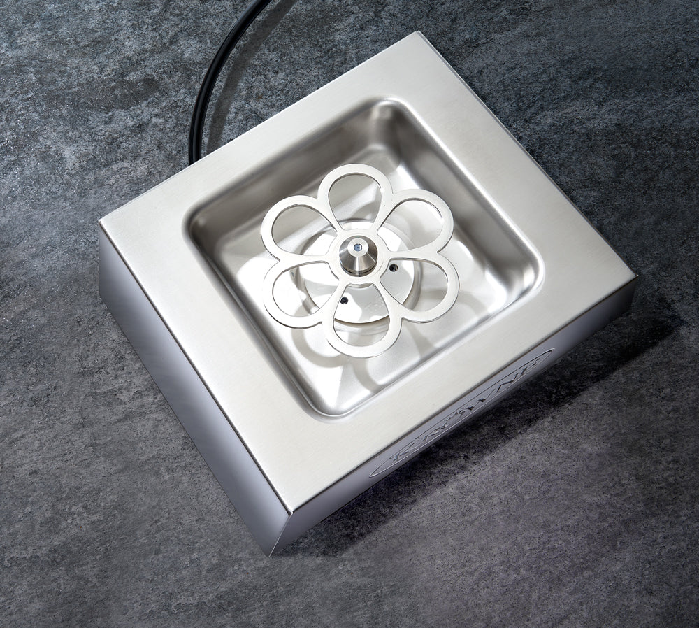 A stainless steel Krowne Royal Series glass chiller with a square basin and a flower-shaped rotating component in the center, placed on a textured gray surface. An electrical cord extends from the top of this clean, modern device.