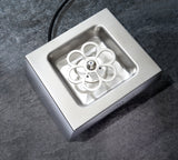 A stainless steel Krowne Royal Series glass chiller with a square basin and a flower-shaped rotating component in the center, placed on a textured gray surface. An electrical cord extends from the top of this clean, modern device.