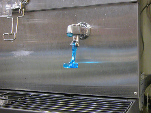 A Scotsman metal beverage dispenser with a spigot is dispensing a stream of bright blue liquid. The liquid drips toward a black grate below, with blue residue visible on the spigot and nearby surfaces. Ideal for use with an ID150 water glass filler kit.