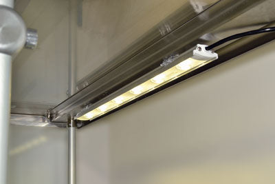 A close-up view of a mounted LED strip light fixed under a metallic shelf or cabinet, emitting bright warm white light. Electrical wires are visible connected to the fixture, with a smooth wall and stainless-top food shields nearby.