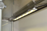 A close-up view of a mounted LED strip light fixed under a metallic shelf or cabinet, emitting bright warm white light. Electrical wires are visible connected to the fixture, with a smooth wall and stainless-top food shields nearby.