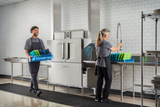 Two people in gray uniforms work in a commercial kitchen: one carries a blue rack of trays, smiling, while the other rinses trays before loading them into a conveyor dishwasher. The background is white tiled walls and shelves with clean plates.