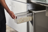 A person’s hand pulls a metal basket filled with shredded vegetables from a stainless steel Hobart CL66VL-3 appliance. The shiny machinery and food preparation setting, equipped with ventless energy recovery, suggest a professional kitchen environment.