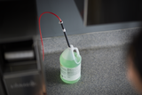 A translucent gallon jug filled with green liquid sits on a speckled gray countertop. A black and white dispensing tube with a red hose, often found near a conveyor dishwasher, is inserted into the jug’s opening. The background includes a gray wall and stainless steel appliance.