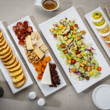 Four rectangular melamine dishes on a beige tablecloth display toasted bread slices, assorted cured meats and cheeses with nuts and dried fruit, a mixed green salad, and rows of tomatoes with mozzarella and basil. All dishes are dishwasher safe.
