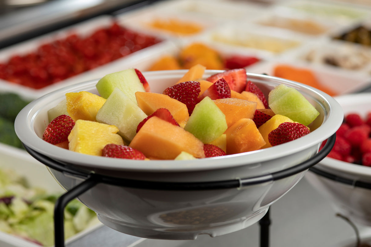 A white serving bowl filled with assorted fresh fruit—strawberries, cantaloupe, honeydew, and pineapple chunks—sits on a metal stand. In the background, trays of other fruits and salad ingredients are slightly out of focus.