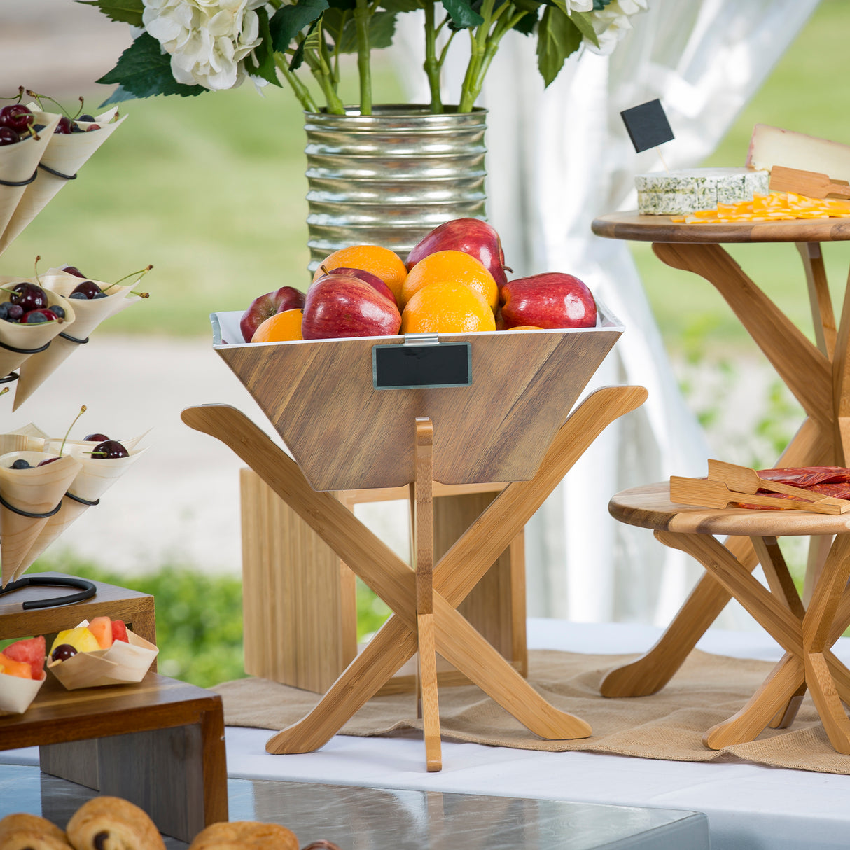 A wooden fruit bowl with acacia wood finish holds apples and oranges on a stand atop a white table. Nearby are trays with cheese, fruit cups, pastries, and a metal vase of white flowers in the background. The setting appears to be outdoors.