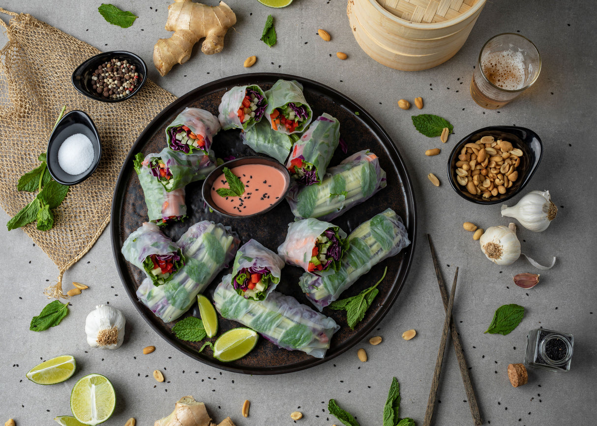 A round tray with fresh vegetable spring rolls, halved and arranged around a pink dipping sauce in a dishwasher safe Frostone Collection Bowl. Surrounding the tray are garlic bulbs, peanuts, ginger, herbs, lime wedges, spices, and chopsticks on a gray surface.