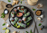 A round tray with fresh vegetable spring rolls, halved and arranged around a pink dipping sauce in a dishwasher safe Frostone Collection Bowl. Surrounding the tray are garlic bulbs, peanuts, ginger, herbs, lime wedges, spices, and chopsticks on a gray surface.