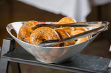A bowl from the Dalton II Collection holds assorted bagels, with a stainless steel tong server resting on top. The bowl sits on a dark tray, against a blurred background and white tablecloth. All items are dishwasher safe for easy cleanup.