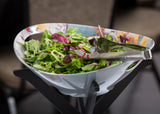A bowl of mixed green salad with leafy greens and purple lettuce sits on a stand. The Dalton II Collection stainless steel tong server rests inside the decorative bowl, ready for serving. A blurred brown chair keeps focus on the fresh salad.