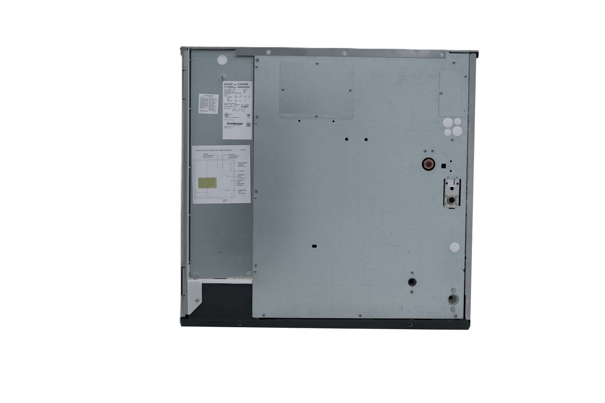 The image shows the back panel of a large metallic appliance, likely a commercial ice machine or dishwasher, with various screws, labels, a circular component, an exposed wiring port, and attached paper documents. The panel is mostly flat with some indents.