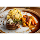 A plated meal featuring a bacon-wrapped filet mignon cooked medium well and topped with herb butter and a bamboo meat marker pick, creamy mashed potatoes with cracked black pepper, colorful glazed baby carrots, and a rich brown sauce.