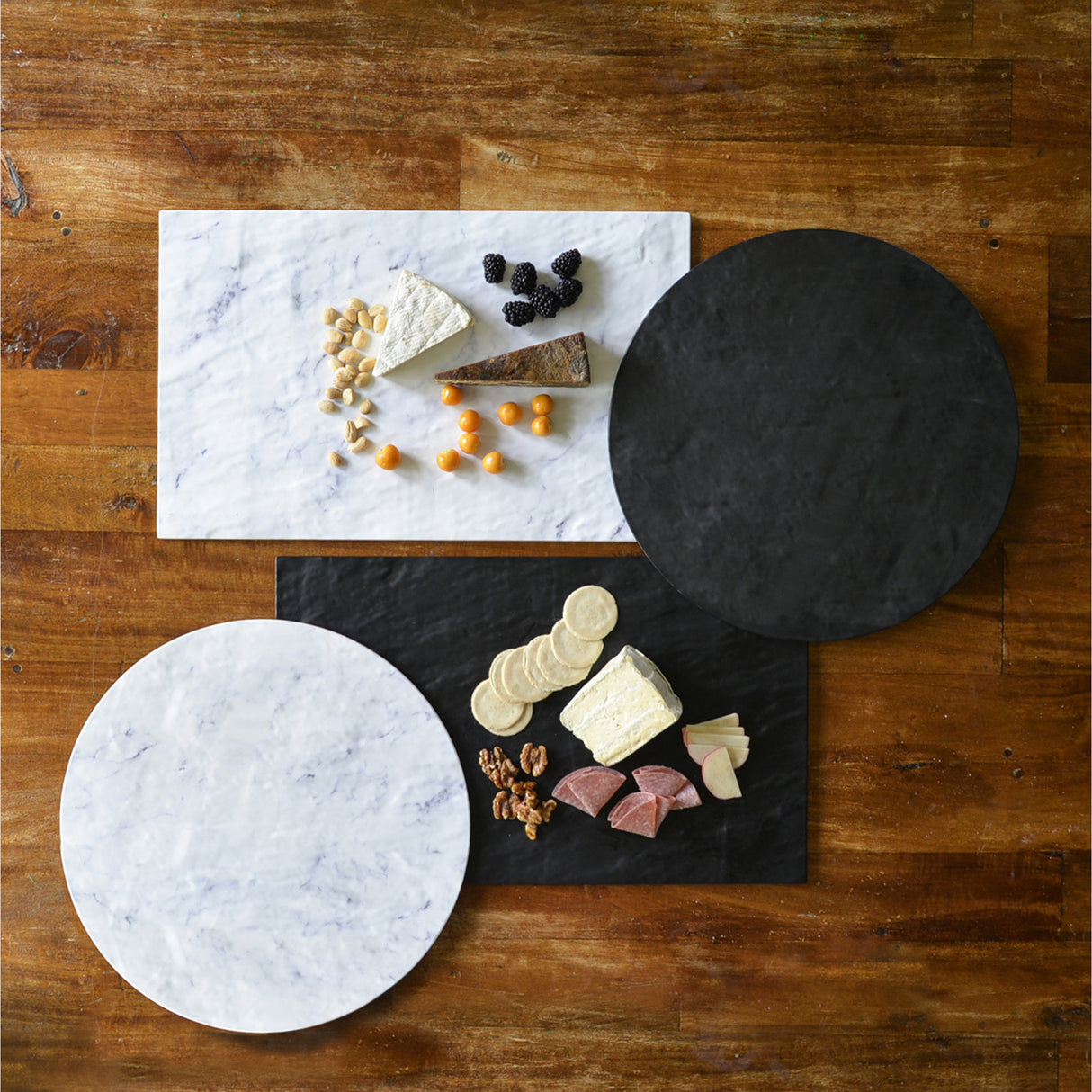 Two rectangular and two round white marble serving boards rest on a wooden table; one is topped with cheese, crackers, apple slices, walnuts, and salami, another with cheese, nuts, blackberries, and orange berries; other boards are empty.