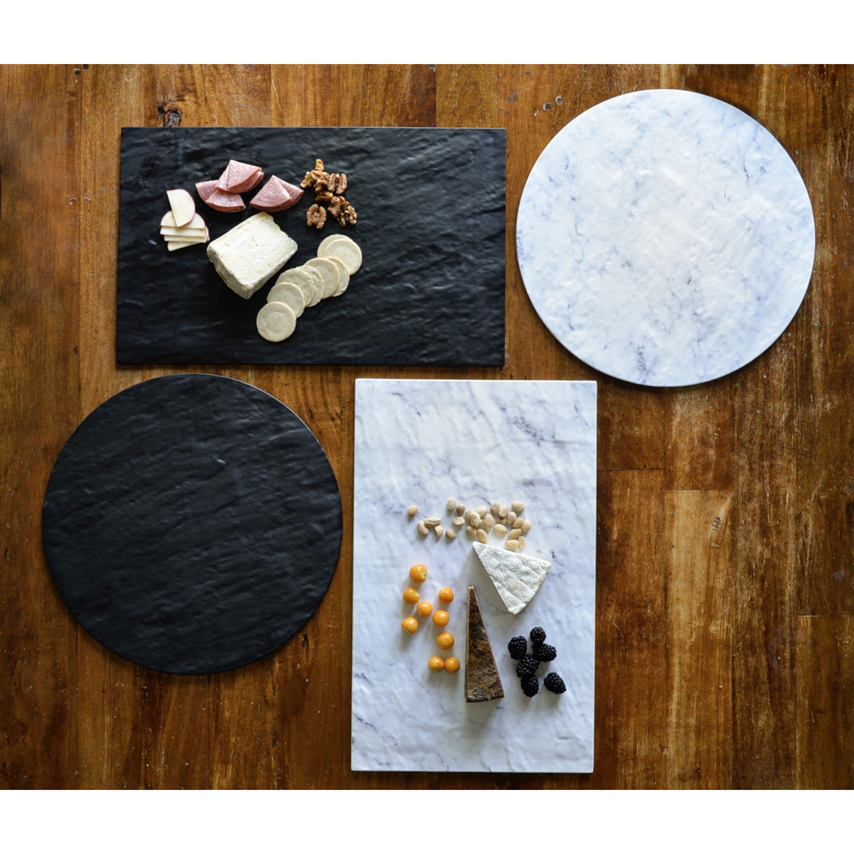 Four marble serving boards—two rectangular, two round—are arranged on a wooden table. Some display cheese, crackers, and fruit, while others simply highlight the contrasting black and white marble surfaces of the Frostone Collection Display Tray.
