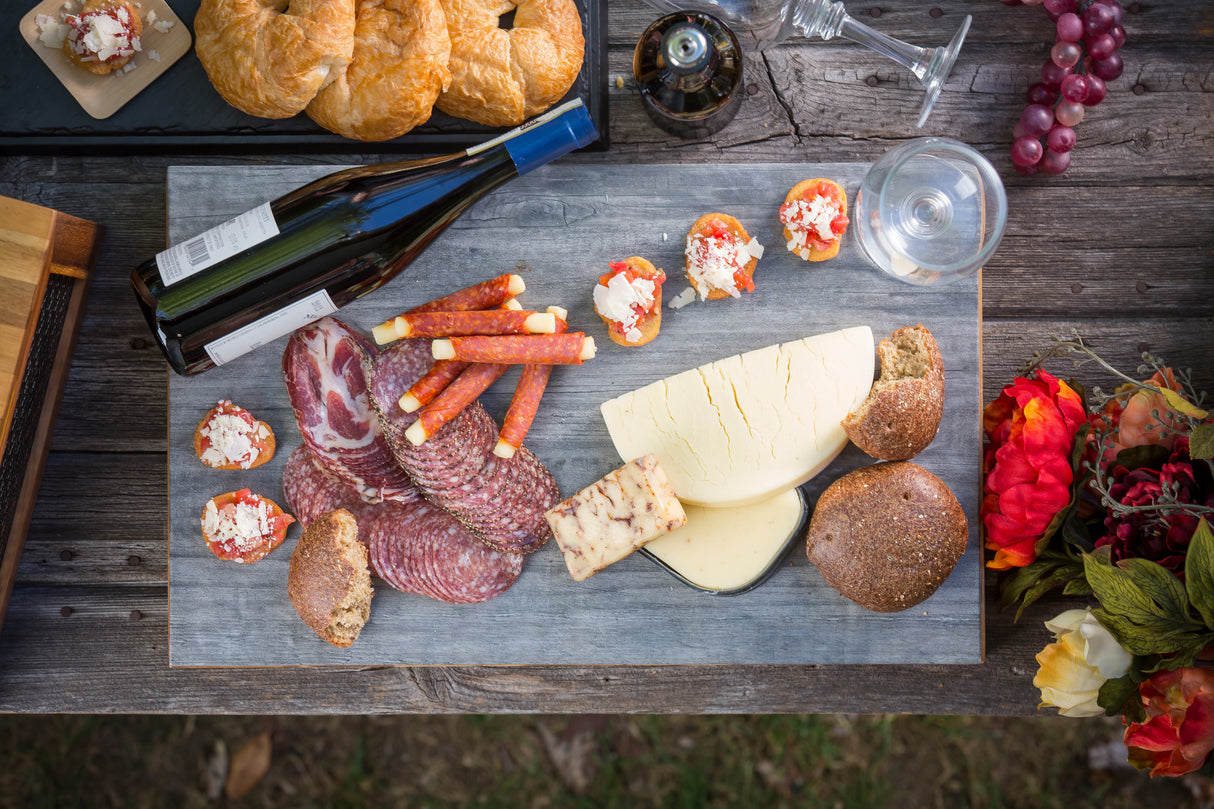 A wooden table displays a charcuterie board with cured meats, cheeses, bread rolls, and bruschetta atop a distressed wood tray. Surrounded by wine, croissants, grapes, and vibrant flowers, it creates a charming rustic picnic setting.