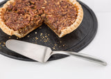 A pecan pie with a slice missing sits on a black tray. An 18/8 stainless steel Buffet Cake Server from the Dalton Collection rests in front of the pie, which has a golden crust and whole pecans atop its rich filling. Crumbs are scattered on the tray.